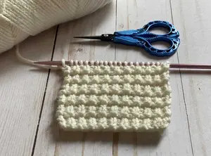 The grid knit stitch pattern swatch worked in white shown with yarn and a pair of scissors