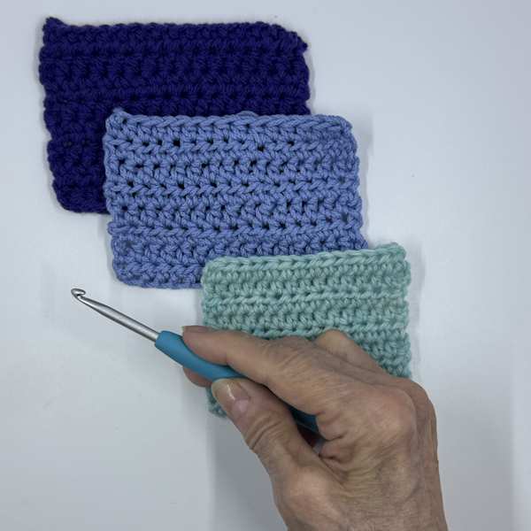 The Clover Amour Ergonomic Crochet Hook is held in front of three crocheted swatches