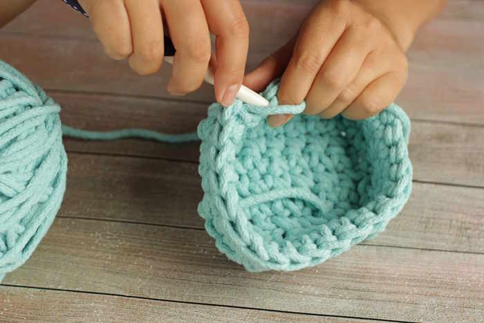 A woman is crocheting a small basket in a light teal colored yarn. A woman is crocheting a small basket in a light teal colored yarn.