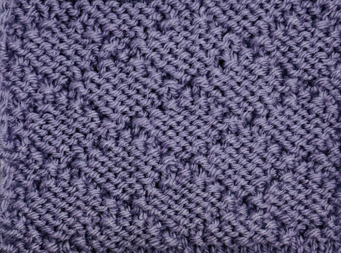 The wrong side is showing for the diamond brocade stitch worked in a purple yarn