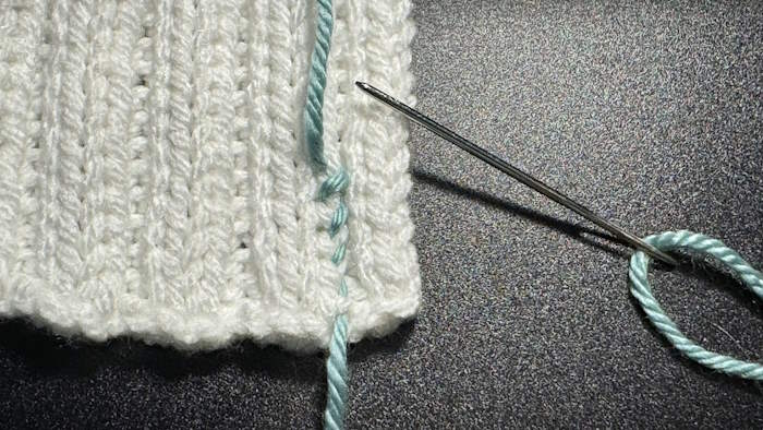 Step one of weaving in ends in rib stitches showing how to insert the yarn needle around each rib stitch from right to left up the column of stitches.