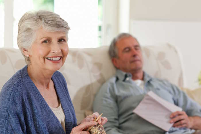 An older woman is knitting while her husband is sleeping